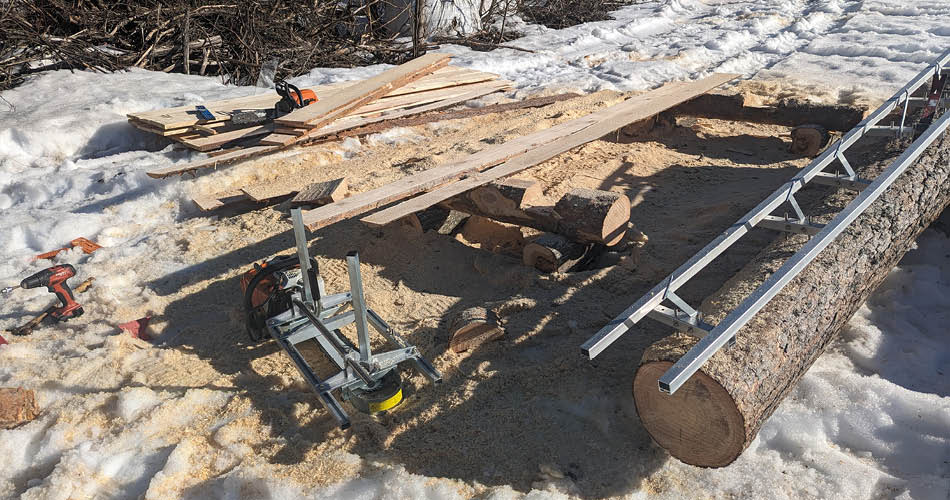 Mini sagbruk 50cm 9064330 med 7 fot skinner i snøen En link til underkategori sagbruk. Bildet viser ei stokke med montert skinner på. Foran stokken er et arbeidsbenk laget av to stokker med ferdig kledning på. Det er snø på bakken og spredt rundt er ferdig reisverk, en motorsag, en drill og mini sagbruket.