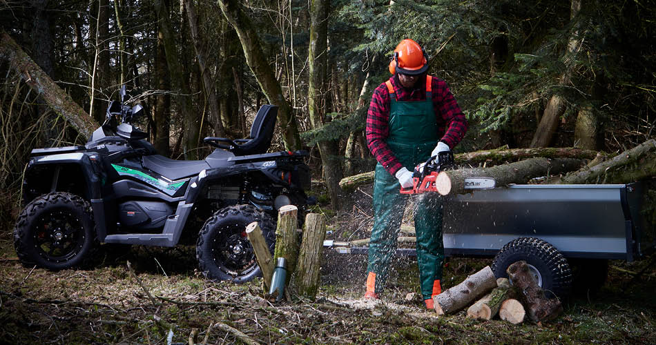 Vedhåndtering med Odes ATV, henger med tipp 9036232 og verneutstyr En link til underkategori sikkerhetsutstyr. Bildet viser en mann i full verneutstyr som kapper ei stokke i ringer foran en ATV-henger. En Odes ATV står foran en skog med ved og termosflaske også vist